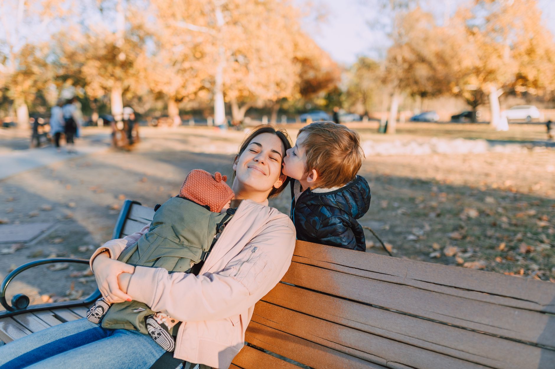 boy kissing her mom sitting on brown wooden bench carrying a baby